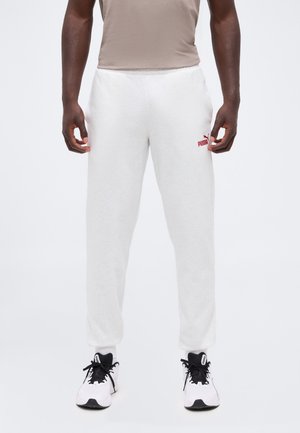 Homme portant un pantalon de survêtement blanc Puma, une chemise beige et des baskets blanches avec des lacets noirs, debout sur un fond blanc.