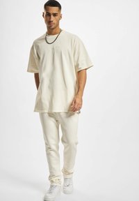 Lys beige oversized T-skjorte med brodert tekst, kombinert med matchende tettsittende bukser. Modellen bruker hvite joggesko. Glatt bomullstekstur.