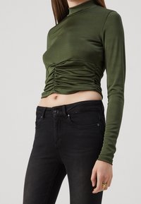 Top de manga larga verde oliva con cuello alto y detalle fruncido en la cintura, combinado con jeans negros ajustados. Textura de tela suave.