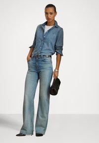 Gaiši zils džinsa krekls ar pogām, garām piedurknēm. Plaša piegriezuma izbalējušas zils džinsi. Melna somiņa ar liektas formas, ko nēsā modele.