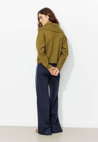 Pull court vert olive avec un large col, associé à un pantalon marine avec des rayures bleues sur les côtés et une coupe décontractée, se tenant devant un mur blanc.