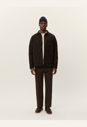 Black wool jacket with button closure and front pockets, paired with a cream sweater, brown pants, and a blue knit beanie.