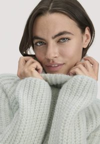 Frau mit grünen Augen, die den Kragen eines hellgrauen, grob gestrickten Pullovers nah an ihr Gesicht hält, vor einem neutralen Hintergrund.