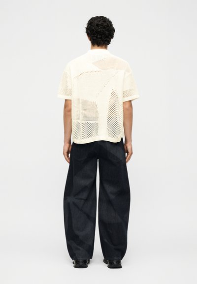 Άνδρας με σγουρά μαλλιά που φοράει χαλαρό, κρεμ χρώματος κοντομάνικο πουκάμισο από δίχτυ και φαρδιά σκούρα τζιν παντελόνια, από πίσω σε μονόχρωμο φόντο.