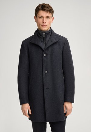 Mann trägt einen dunkelgrauen, geknöpften Wollmantel über einer schwarzen Reißverschlussjacke und steht vor einem einfarbigen hellgrauen Hintergrund.