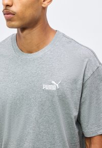Homme portant un T-shirt Puma gris clair à col rond avec un logo Puma blanc sur la poitrine gauche, sur un fond clair uni.