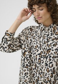 Halenka s leopardím vzorom v čiernej, hnedej a krémovej farbe, s klasikou golierom, predným zapínaním na gombíky a dlhými rukávmi so voľným strihom.
