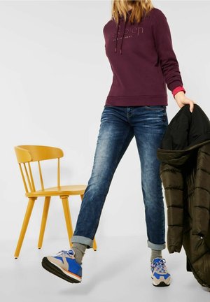 Dark purple hoodie with logo, blue jeans, grey socks, and blue sneakers. Black puffer jacket held in one hand. Yellow chair in background.