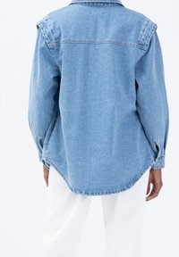 Chemise overshirt en denim bleu clair avec des manches longues, un col pointu et des poignets boutonnés. Présente un ourlet arrondi et des détails de couture visibles.