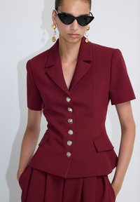 Donna che indossa un blazer bordeaux aderente con sei bottoni decorativi, una gonna coordinata plissettata, occhiali da sole a forma di gatto neri e orecchini a pendente dorati.