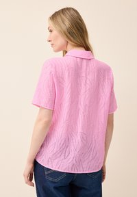 Blusa rosa a maniche corte con un motivo a onde testurizzate, con colletto e orlo curvo, abbinata a pantaloni di denim scuro.