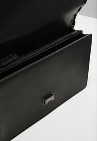 Černá kožená kabelka s otevřeným víkem, která odhaluje vnitřní zipovou přihrádku a stříbrný obdélníkový uzávěr na přední straně.