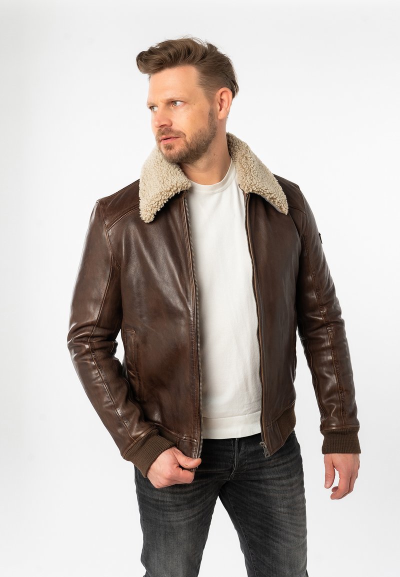 Giacca bomber in pelle marrone con colletto in shearling beige, polsini a costine e tasche laterali, indossata sopra una t-shirt bianca e jeans scuri.
