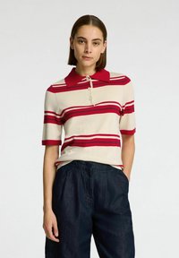 Gebreide poloshirt in crèmekleur met rode strepen, voorzien van een rode kraag en drie knopen sluiting. Gecombineerd met donkergekleurde, geplooide wijde broek.