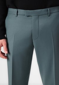 Pantalones de color verde pizarra a medida con una textura suave, frente plano y un bolsillo lateral discreto, con trabillas para el cinturón en la cintura.