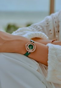 Reloj con correa de cuero verde y caja redonda de oro rosa, embellecida con cristales, y una esfera verde con marcadores de hora plateados.