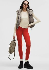 Chaleco gris acolchado sobre una blusa beige clara, leggins rojos y botas negras con puños texturizados. La modelo sostiene un bolso suave gris.