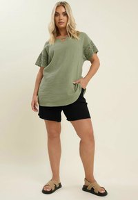 Olivgrön kortärmad topp med nyckelhålsdetalj och spetsaccenter; kombinerad med svarta shorts och beiga sandaler. Mjuk, texturerad tyg.