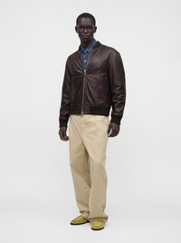 Homme portant une veste bomber en cuir marron foncé sur une chemise bleue, un pantalon beige à jambes larges et des mocassins jaunes, debout sur fond blanc.