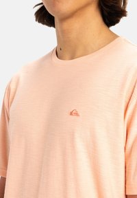 Camiseta de manga corta de color rosa claro, hecha de una tela suave. Presenta un pequeño logo bordado en una tonalidad ligeramente más oscura en el lado izquierdo del pecho.