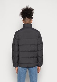 Person med lockigt hår som bär en svart pufferjacka och blå jeans, sedd bakifrån mot en ljus vit bakgrund.