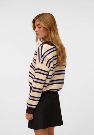Kvinde med langt blondt hår iført en cremefarvet sweater med sorte og lilla striber og et sort skørt, stående i profil med lukkede øjne.
