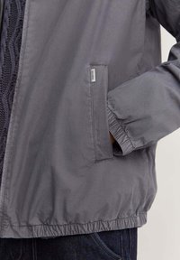 Veste grise légère avec des poignets élastiques et une poche frontale cachée ; texture lisse et coupe décontractée, arborant une étiquette de marque discrète.