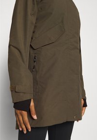 Vue de profil droit d'une personne portant une veste vert olive avec poche zippée, poche à rabat et sangle de poignet réglable sur une manche noire.