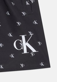 Μαύρο ύφασμα με πολλαπλά λευκά λογότυπα "cK" σε διάφορα μεγέθη και προσανατολισμούς, με κάθετη ραφή κοντά στην κορυφή.