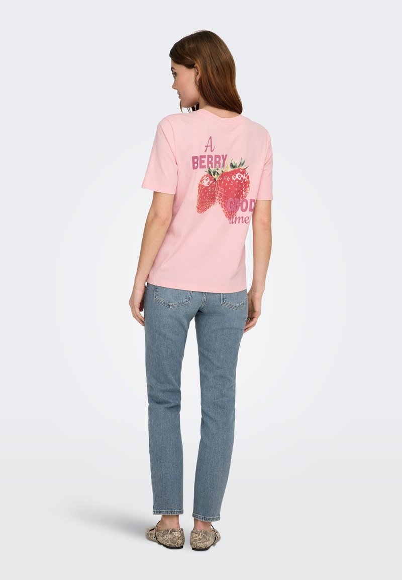 Kvinde med langt brunt hår iført en lyserød T-shirt med jordbær og tekst, blå jeans og mønstrede sko, stående med ryggen mod.