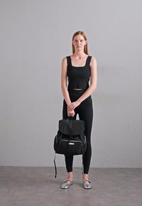 Black backpack with a roll-top closure, contrast white trim, and multiple pockets. Model wears a black fitted crop top and leggings. Silver shoes.
