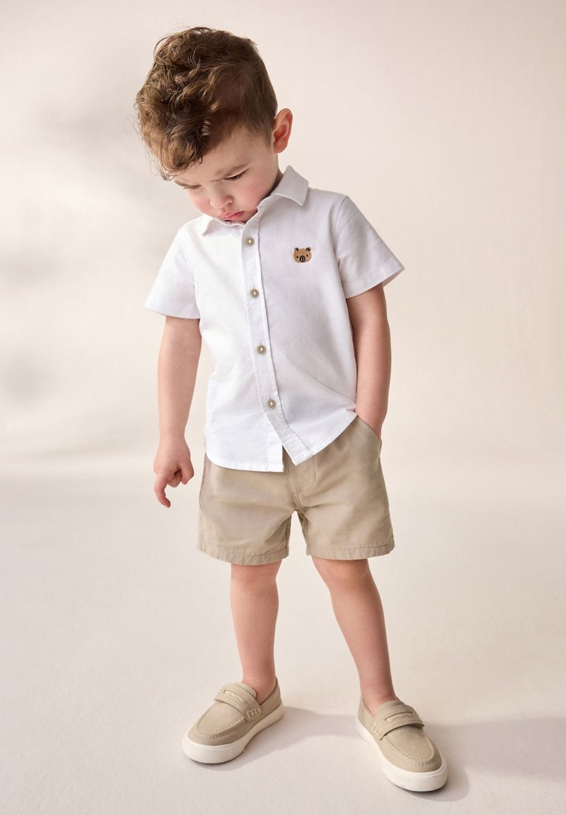 Jeune garçon portant une chemise blanche à manches courtes avec un patch animalier, un short beige et des chaussures beige à enfiler, regardant vers le bas.