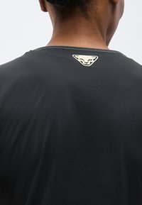 Πλάγια όψη από πίσω ενός ατόμου που φοράει μαύρο πουκάμισο με ένα ανοιχτόχρωμο, στυλιζαρισμένο λογότυπο πανθήρα στο κέντρο κάτω από τον λαιμό.