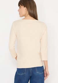 Beige three-quarter sleeve top made of soft fabric with a round neckline. Paired with blue denim bottoms, showcasing a casual fit.
