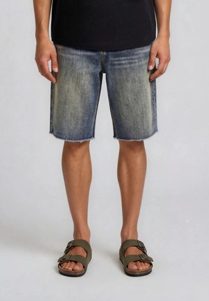 Hombre con pantalones cortos de mezclilla desgastada y sandalias oliva con hebillas, de pie frente a un fondo neutro.