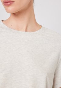 Naise alumine nägu ja kael, kandes heledat halli ümara kaelusega särki ühevärvilise tausta ees.