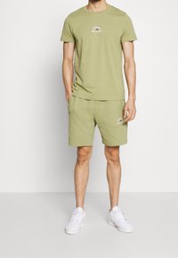 Conjunto de camiseta y pantalones cortos en verde oliva claro. La camiseta presenta un cuello redondo y un pequeño diseño de logo. Los pantalones cortos tienen bolsillos y puños acanalados. Zapatillas blancas.