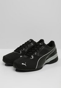 Mustat urheilukengät, joissa on teksturoitu synteettinen päällinen, nauhakiinnitys ja harmaa korostusviiva. Sivussa on valkoinen logo.