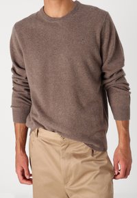 Brązowy sweter z dzianiny wykonany z miękkiego materiału, z okrągłym dekoltem, ściągaczami przy rękawach i subtelnym logo na piersi. W zestawie z beżowymi spodniami.