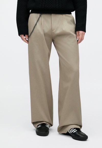 Asmuo, dėvintis smėlio spalvos plačias kelnes su sidabrine grandine, juodą megztinį ir juodus sportbačius su baltais dryžiais.