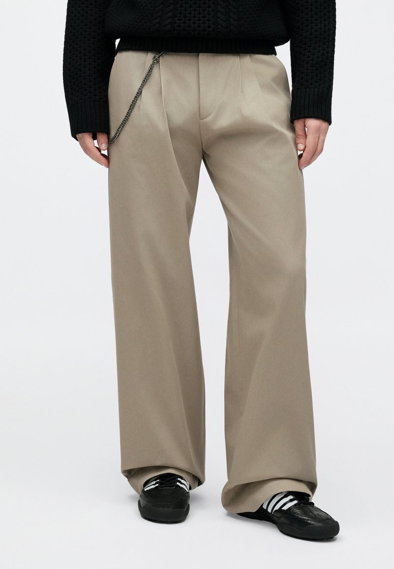 Person wearing beige wide-leg pants with a silver chain, black knit sweater, and black sneakers with white stripes.