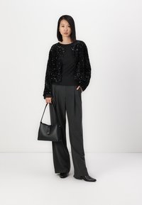 Giacca cropped nera con paillettes, top nero, pantaloni grigi a gamba larga, borsa nera e stivaletti a punta su uno sfondo neutro.