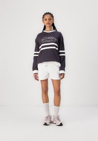Sudadera azul marino oscuro con rayas blancas, logo en la parte delantera; combinada con pantalones cortos blancos. El modelo lleva zapatillas rosas claras y calcetines blancos hasta la altura del tobillo.