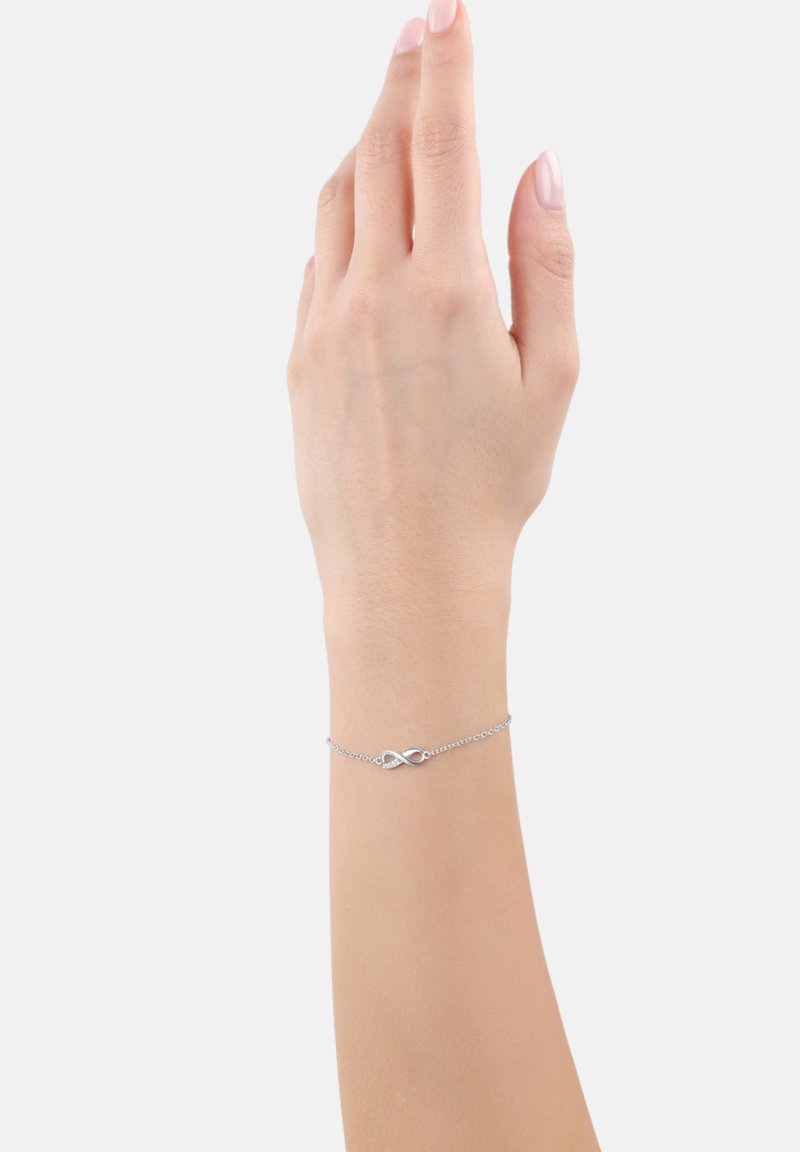 Braccialetto argentato con design a simbolo dell'infinito, caratterizzato da una delicata catena e una superficie liscia e lucida. Indossato su un polso umano.