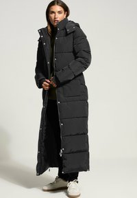 Svart lång pufferjacka med hög krage, tryckknappar och dragkedjestängning. Har en rak silhuett och sydda sektioner för isolering.