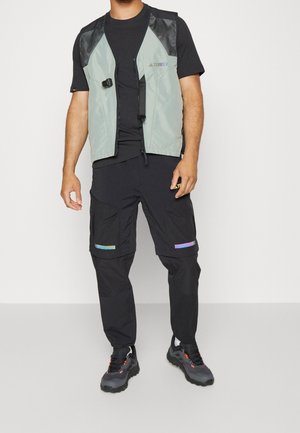 Mies, jolla on päällään vaaleanvihreä ja musta Terrex-liivi mustan t-paidan päällä, mustat cargo-housut heijastinraidoilla ja mustat vaelluskengät.