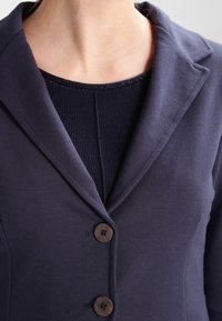 Námořnický modrý blejzr s klopovým límcem, dvěma hnědými knoflíky a texturovaným tmavě modrým topem pod ním. Hladká tkanina se strukturovaným střihem.