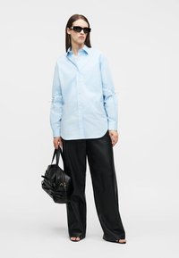 Γυναίκα που φοράει υπερμεγέθη ανοιχτό μπλε πουκάμισο με κουμπιά και φαρδιά μαύρη παντελόνια, κρατώντας μια μαύρη τσάντα χειρός, με γυαλιά ηλίου και ανοιχτά παπούτσια.