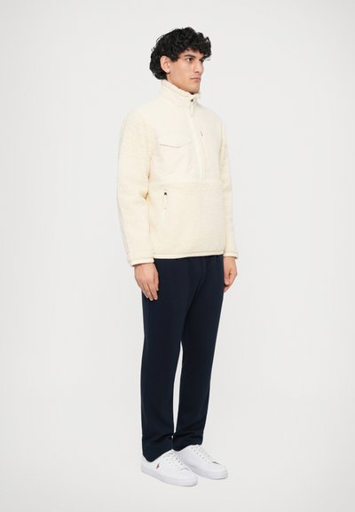Pullover de fleece creme com colar alto, zíper frontal e bolso no peito; combinado com calças de treino azul-escuras e sapatilhas brancas.