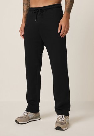 Mand iført sorte sweatpants med lige ben og snøre i taljen samt beige New Balance sneakers, stående på et lyst gulv.